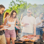 Group of people standing around grill, chatting, drinking and eating.